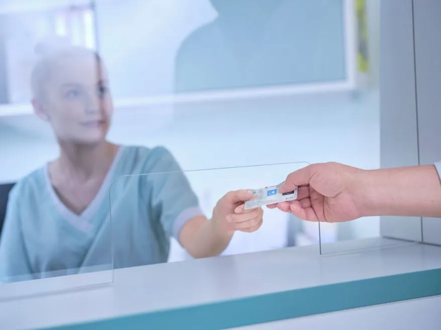 Eine Person übergibt eine Gesundheitskarte an eine Mitarbeiterin an einem Empfangstresen in einer medizinischen Einrichtung.