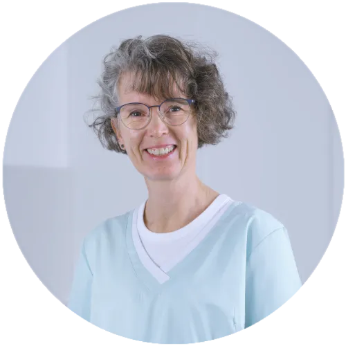 Lächelnde Frau in medizinischer Kleidung mit Brille, kurze lockige Haare, freundlicher Ausdruck.