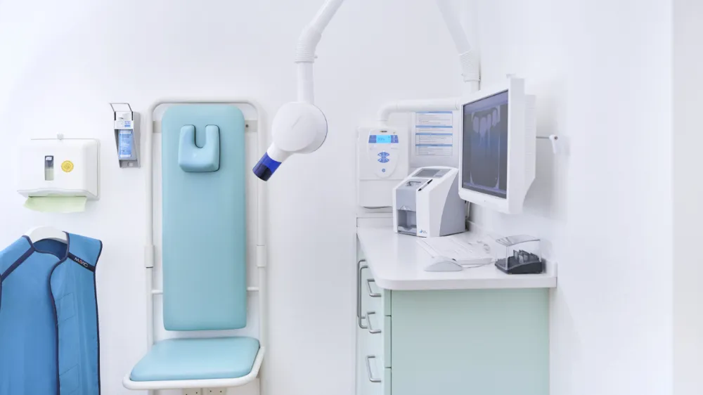 Behandlungsstuhl in einer Zahnarztpraxis mit blauer Polsterung, Röntgengerät und Monitor, auf dem Zahnaufnahmen angezeigt werden.