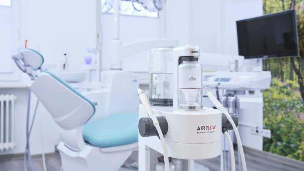 AIRFLOW Prophylaxis Master von EMS, ein Zahnreinigungsgerät mit transparentem Behälter, in einem modernen Zahnarztpraxisraum mit einem Behandlungsstuhl und Monitor im Hintergrund.