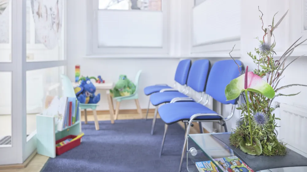 Wartezimmer mit blauen Stühlen, einem Kindertisch und Spielzeug. Auf einem Tisch steht ein Blumenarrangement.