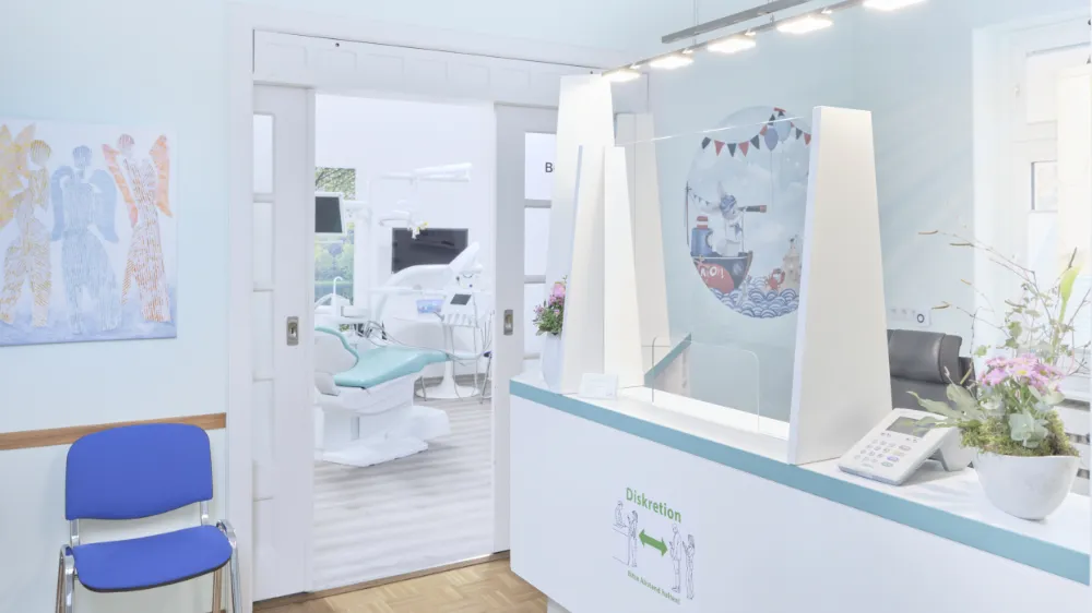 Empfangsbereich einer Zahnarztpraxis mit einem transparenten Sichtschutz, blauer Empfangstheke und einer blauen Wartebank. Im Hintergrund sind Zahnarztstühle und medizinische Geräte sichtbar. An der Wand hängt ein farbenfrohes Kunstwerk.