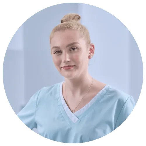 Junge Frau in hellblauer medizinischer Kleidung, lächelnd, mit hochgestecktem Haar. Hintergrund unscharf, professioneller Kontext.