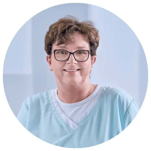 Frau mit Brille in hellblauer Arbeitskleidung, lächelt freundlich in einem medizinischen Umfeld.