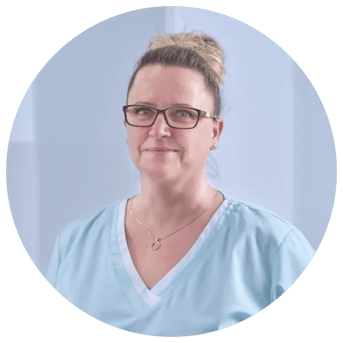 Fachkraft in hellblauer Arbeitskleidung mit Brille, freundlich lächelnd, vor einem hellen Hintergrund.