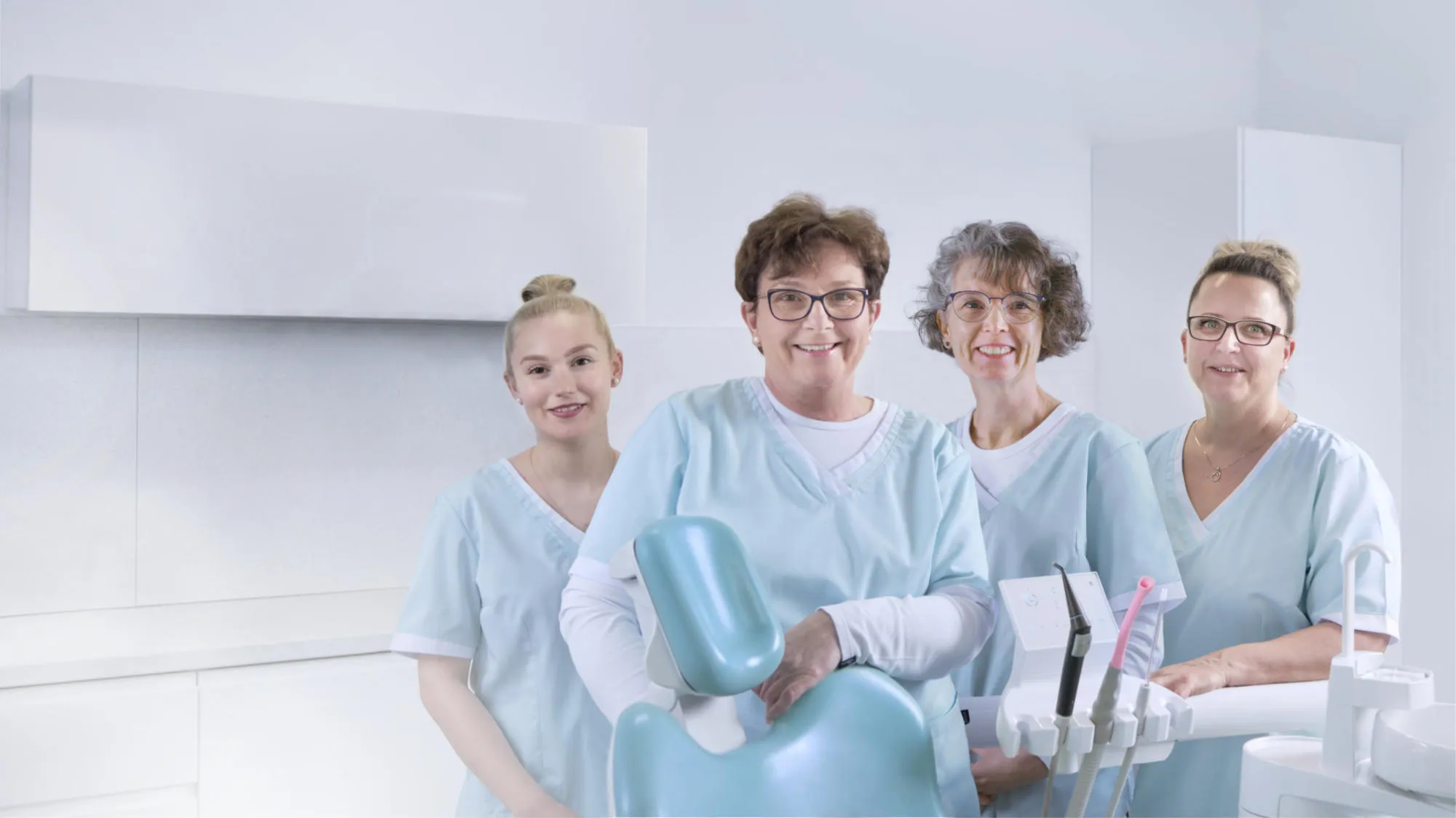 Vier Zahnarzthelferinnen in blauen Kitteln stehen lächelnd vor einem zahnmedizinischen Behandlungsstuhl und Instrumenten in einer modernen Zahnarztpraxis.